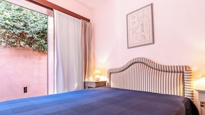 Habitación de hotel con cama, mesita de noche con lámpara, ventana con cortinas e imagen enmarcada en la pared.