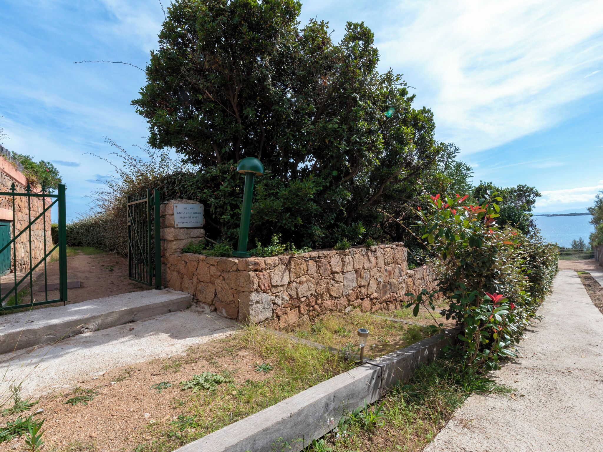 A stone wall with a metal gate, a paved path, and a sign indicating a village.