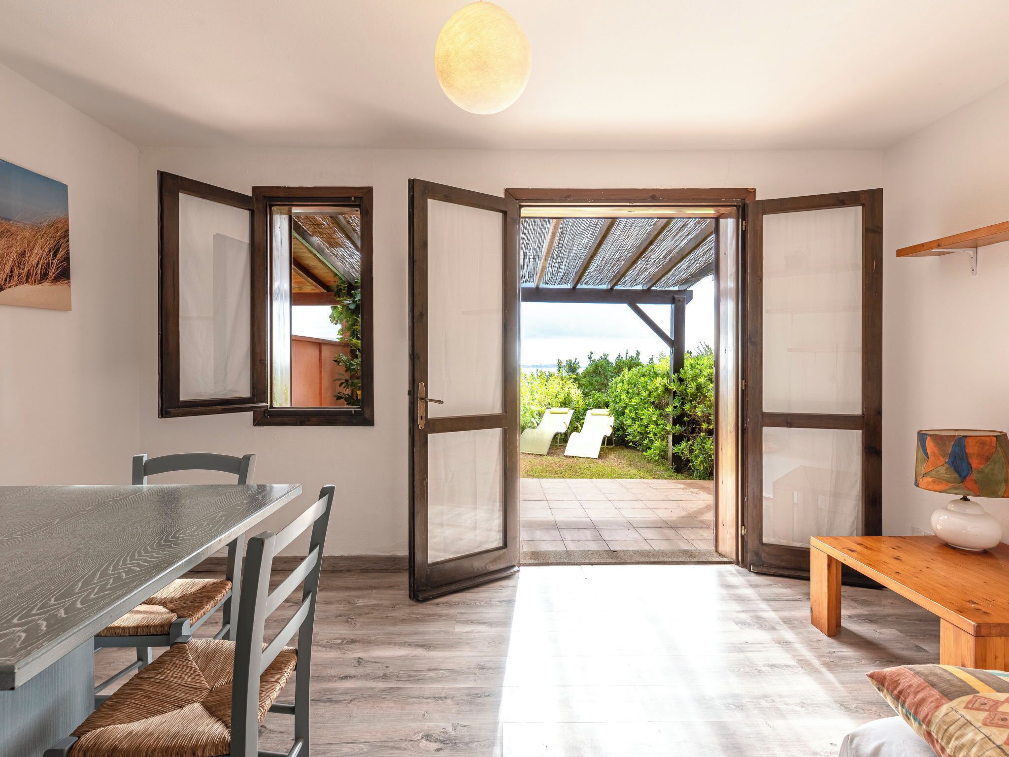 A room with a dining table, chairs, a bench, a sofa, and a view of a garden through open doors.