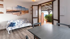 Ein gemütliches Zimmer mit Holzboden, einer Couch mit blumigem Bezug in Blau und Weiß, einem Esstisch mit Stühlen und Blick auf den Strand durch eine offene Tür.