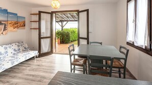 Ein Zimmer mit Holzboden, einem Esstisch mit vier Stühlen, einem Sofa mit Blumenmuster und Blick auf einen Garten durch eine offene Tür.