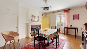 Un espace repas comprenant une table, des chaises, un fauteuil en osier, une cheminée et une vue sur l’extérieur.
