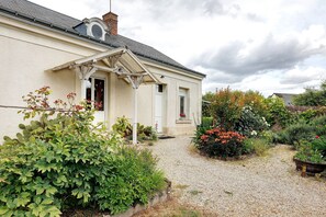 Uma casa com entrada de automóveis em cascalho, um jardim com diversas plantas e flores, e fachada branca.