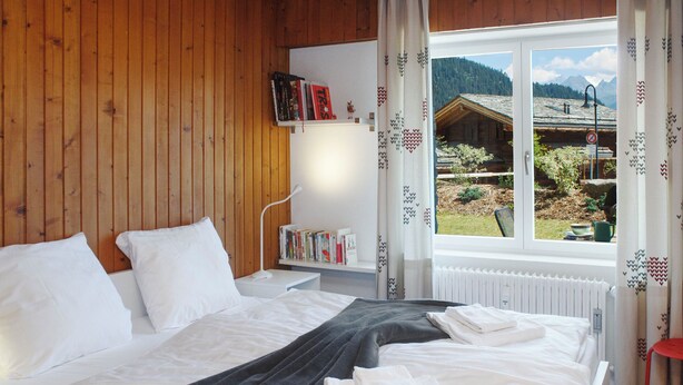 Une chambre à coucher avec un lit, des oreillers, une table de chevet avec des livres, une fenêtre donnant sur une vue de montagne et un radiateur.