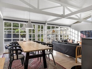 Une pièce spacieuse avec une table à manger en bois, des chaises noires, un canapé gris et un téléviseur. De grandes fenêtres laissent entrer la lumière naturelle et offrent une vue sur la verdure environnante.