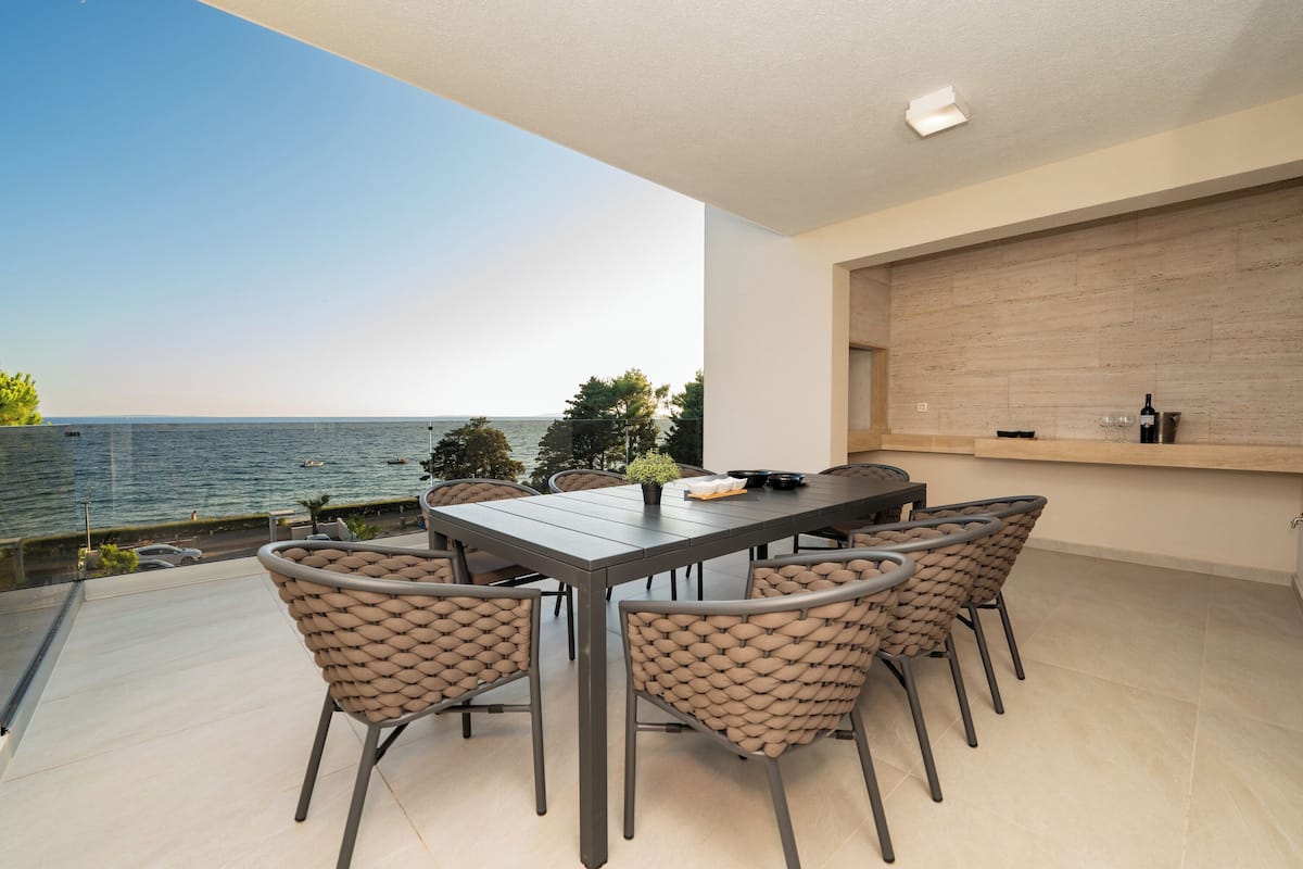 A modern outdoor dining area with a view of the sea, a wooden wall, and a built-in shelf with a bottle and glasses.
