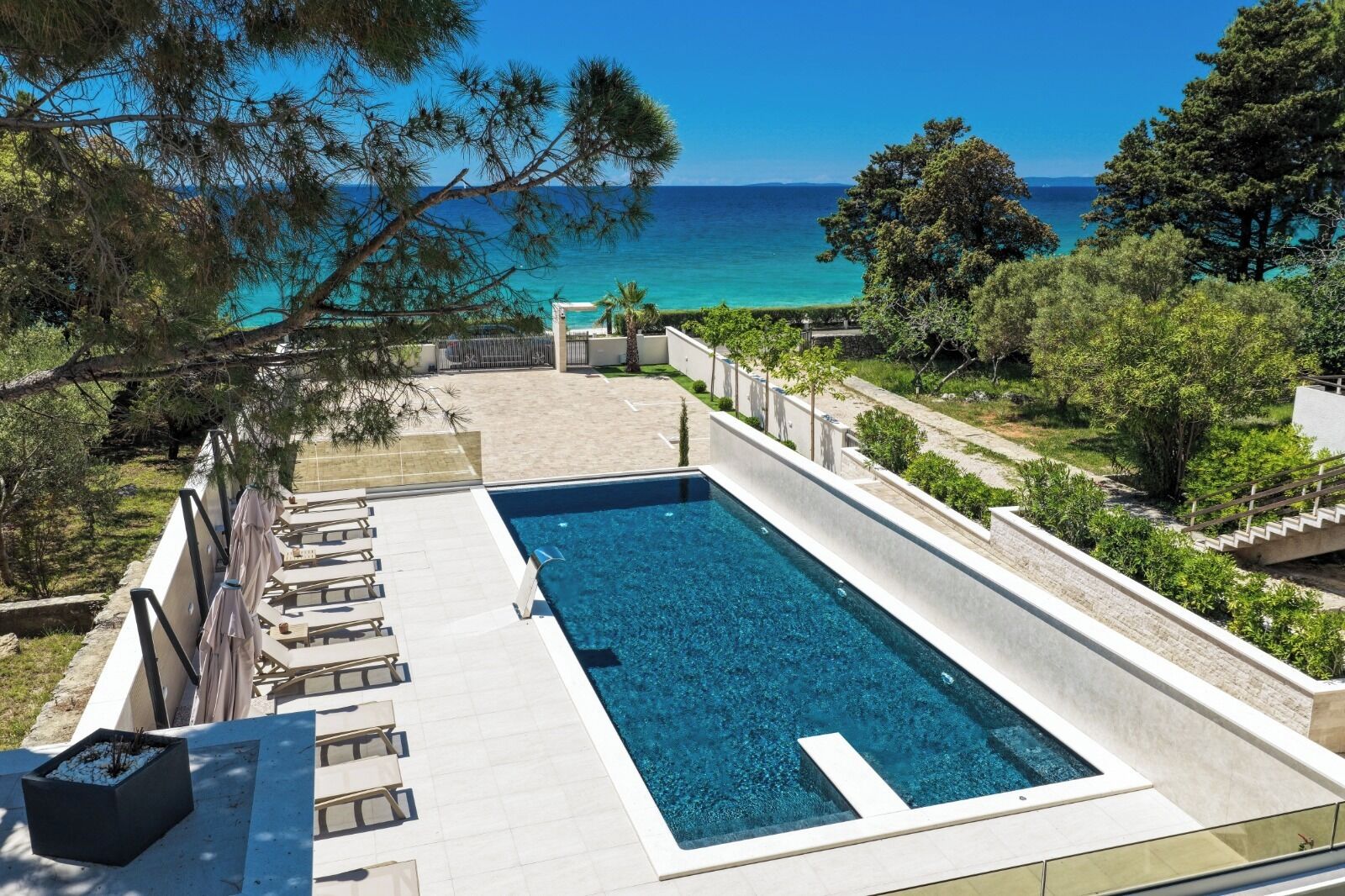 Une piscine rectangulaire à l’eau d’un bleu limpide, entourée d’une terrasse blanche, et offrant une vue sur une plage bordée d’arbres et un ciel dégagé.