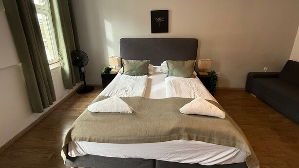 A neatly made bed with a grey headboard, two bedside tables with lamps, a dark sofa, and a window with curtains.