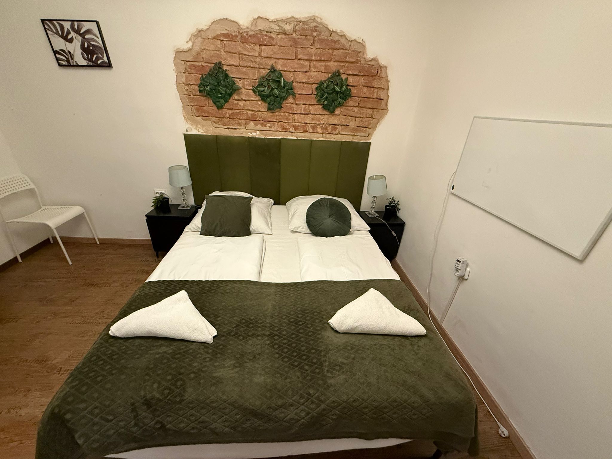 Habitación de hotel con cabecero de pared de ladrillo, dos camas con ropa de cama blanca y verde, una silla blanca y una pequeña mesa con lámparas.
