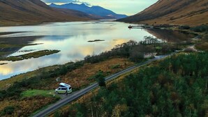 A remote lakeside road with a vehicle and a small building, surrounded by hills and mountains. - Ben Roamer VW Campervan – Explore Scotland in Comfort & Freedom (Dunblane)