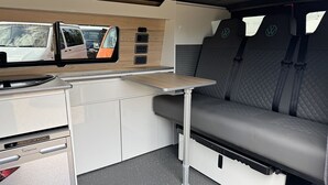 A compact kitchen area with a built-in stove, a small table, and a bench with storage underneath. - Ben Roamer VW Campervan – Explore Scotland in Comfort & Freedom (Dunblane)