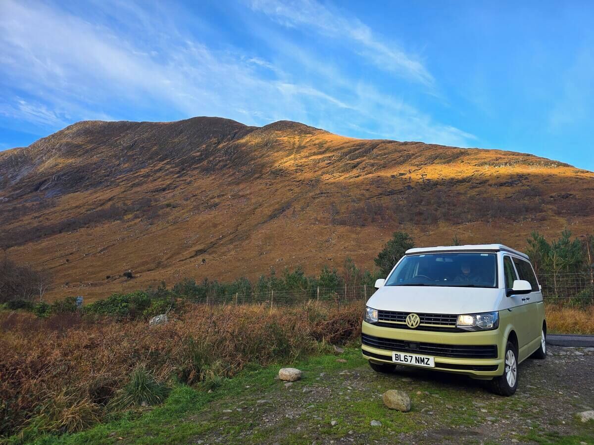 Ein weißer VW-Bus steht auf einer Wiese vor einer Bergkulisse.