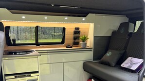 A compact kitchen with a small refrigerator, a sink, and a window with a view of greenery. - Ben Roamer VW Campervan – Explore Scotland in Comfort & Freedom (Dunblane)