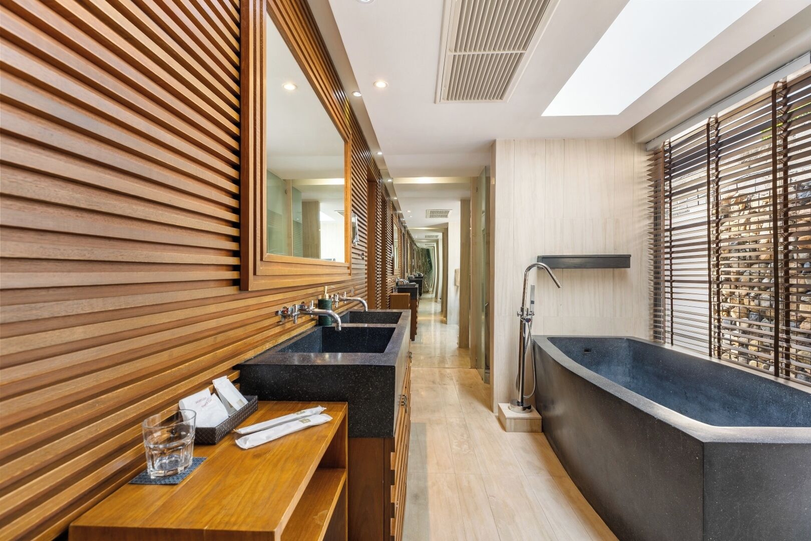 Une salle de bain moderne avec un lambris en bois, un grand miroir, deux vasques, une baignoire et une fenêtre avec des stores.