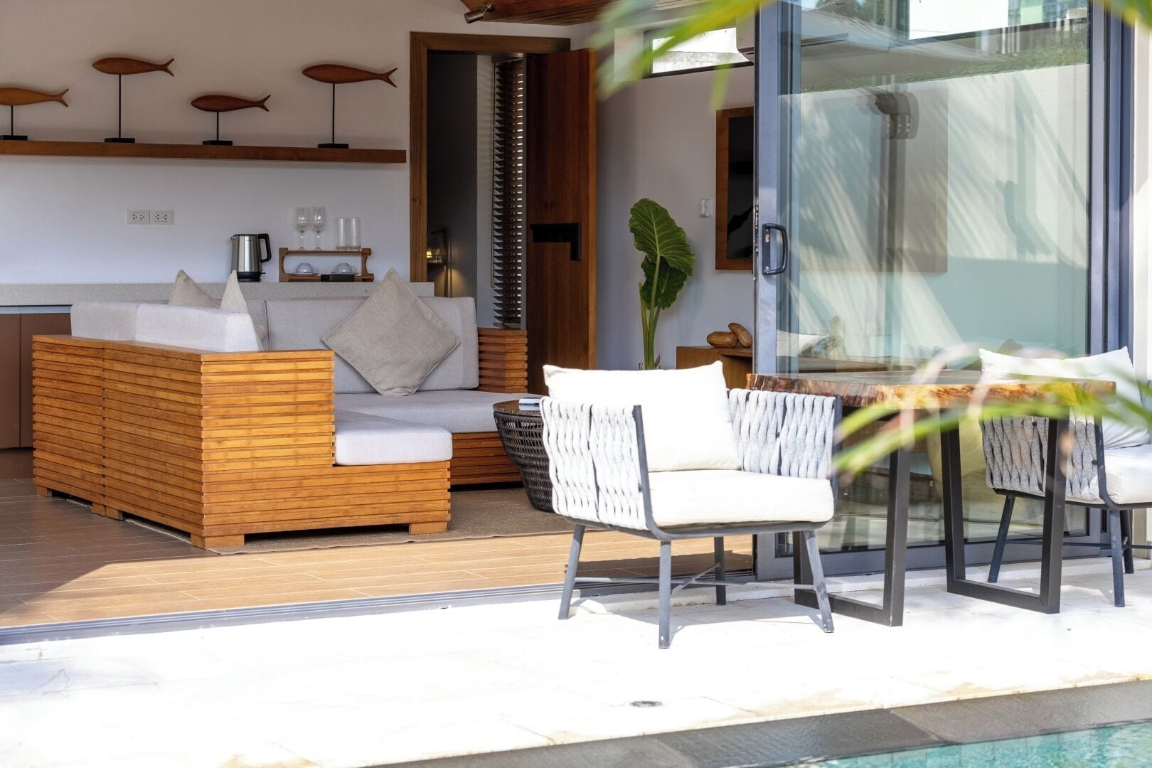 A modern outdoor living area with a wooden sofa, white wicker chairs, a glass table, and a pool.