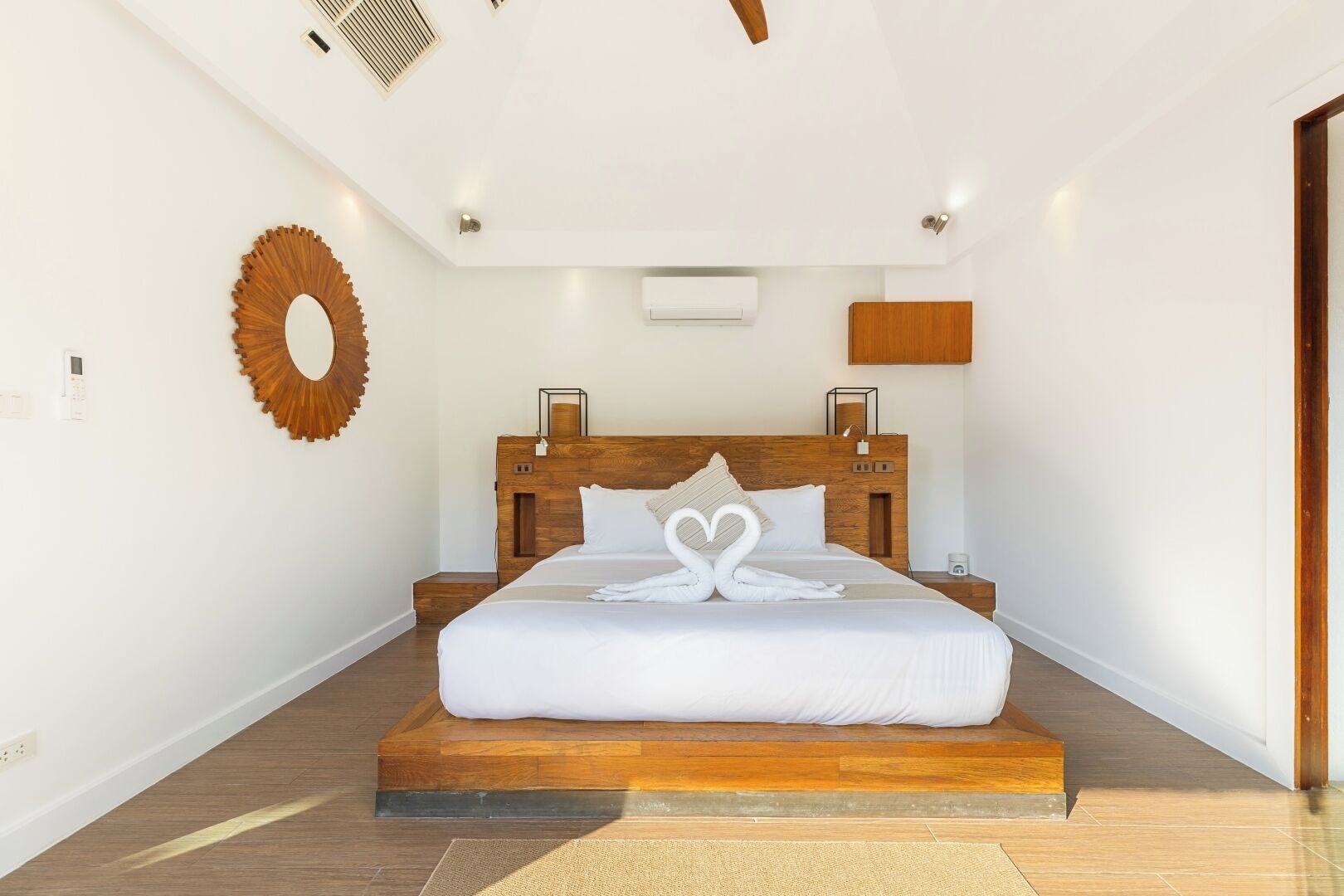 A modern bedroom with a wooden headboard, a large bed, a wall-mounted mirror, and a built-in air conditioning unit.