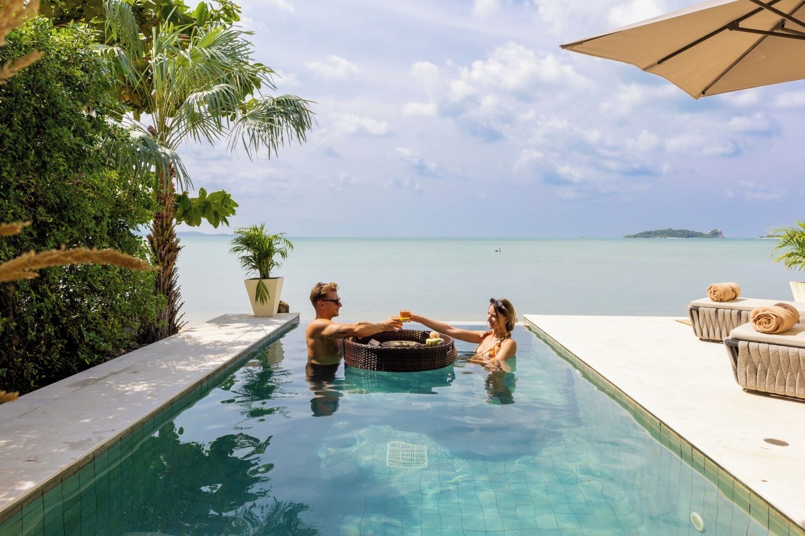 A couple in a clear pool with a view of the sea and an island, surrounded by greenery and a lounge area with a sun umbrella.