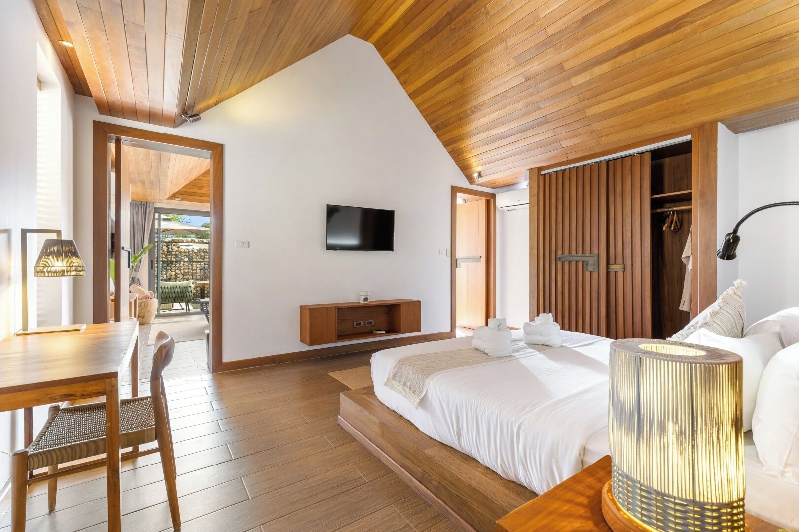 A bedroom with a wooden ceiling, a TV mounted on the wall, a bed with white bedding, a bedside table, a lamp, and a wardrobe.