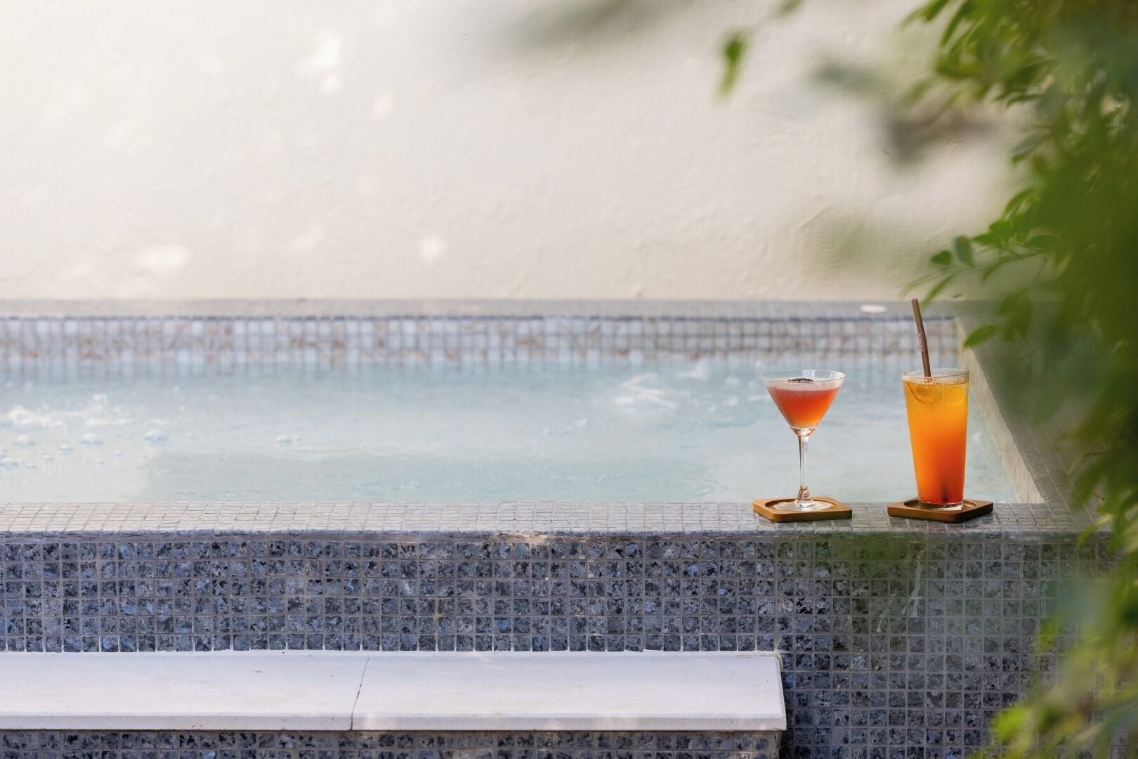 A poolside area with a tiled edge, two drinks on coasters, and a bench.