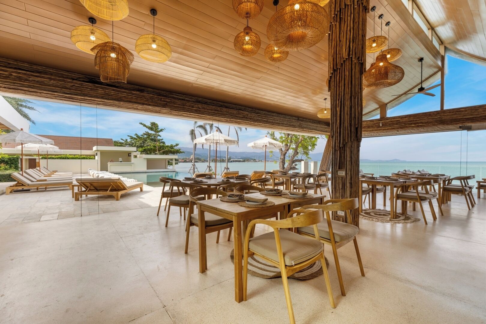 An outdoor dining area with wooden tables and chairs, surrounded by a covered seating area with umbrellas, overlooking a body of water.