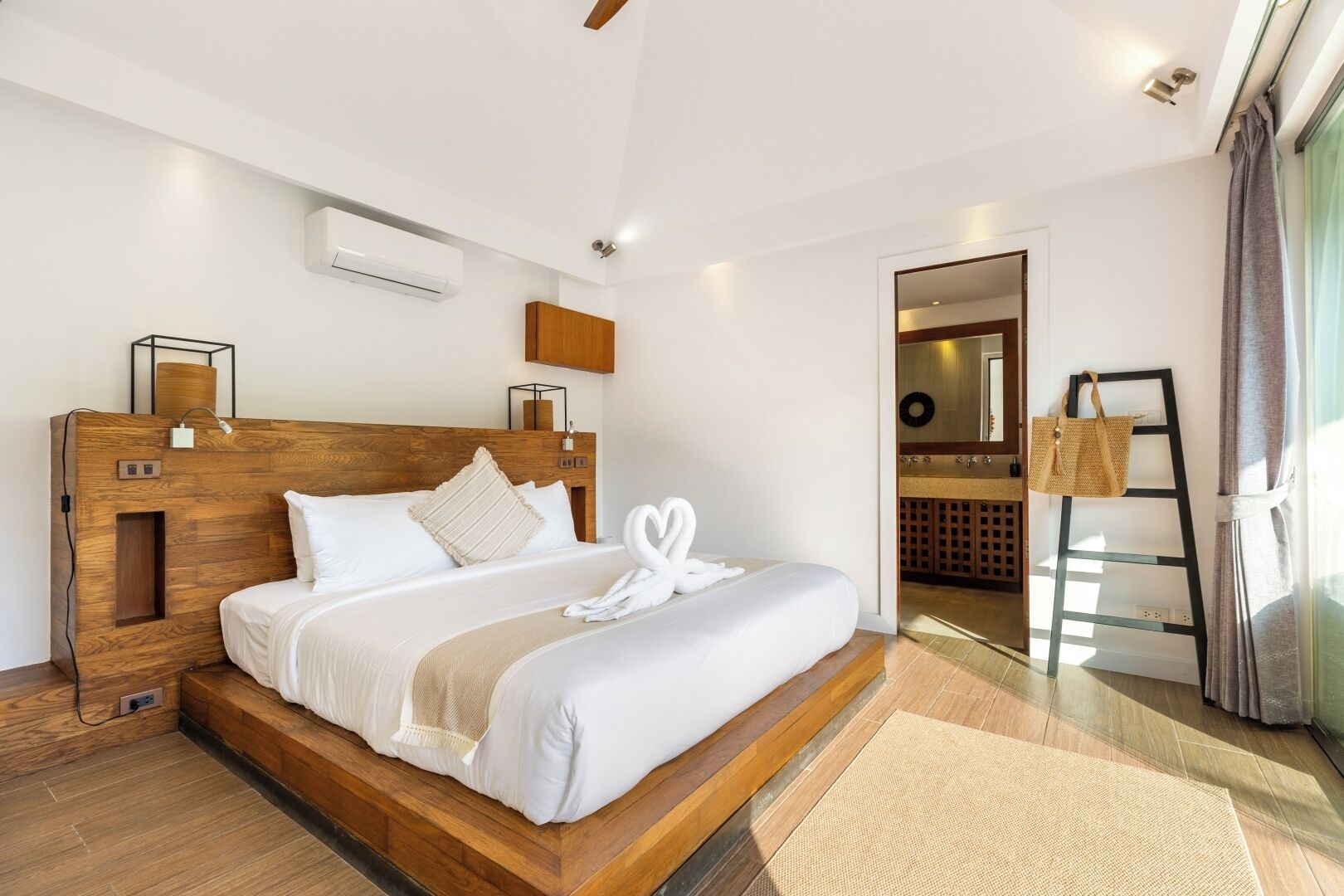 Une chambre d’hôtel moderne avec une tête de lit en bois, un lit recouvert de draps blancs, un grand miroir, une échelle en bois et une vue sur la salle de bain.