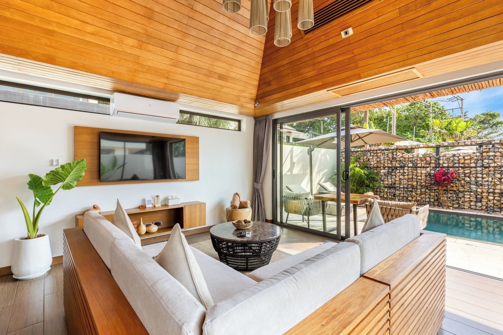 Una sala moderna con techo de madera, un televisor grande montado en la pared, un sofá cómodo, una mesa de centro y vista a un área de piscina al aire libre.