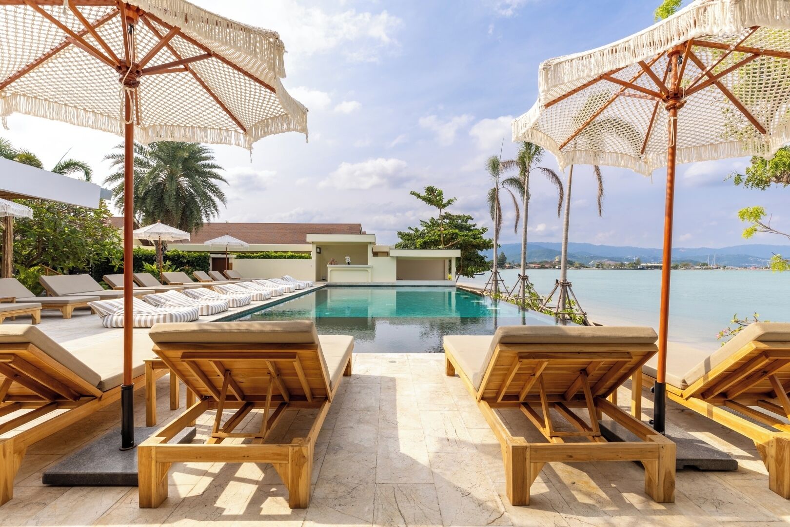 A poolside area with wooden lounge chairs and umbrellas, a clear pool, and a view of the sea and mountains.