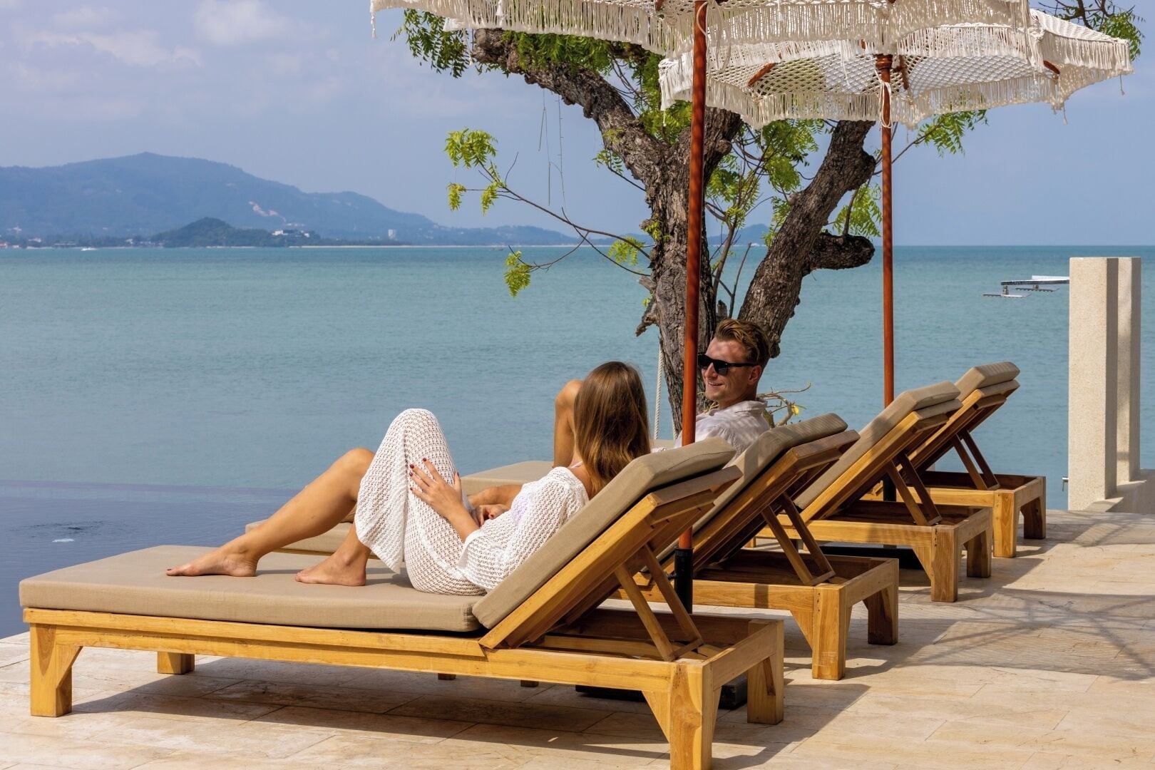 Ein Strandloungebereich mit Holzliegen, einem Baum und Blick aufs Meer.