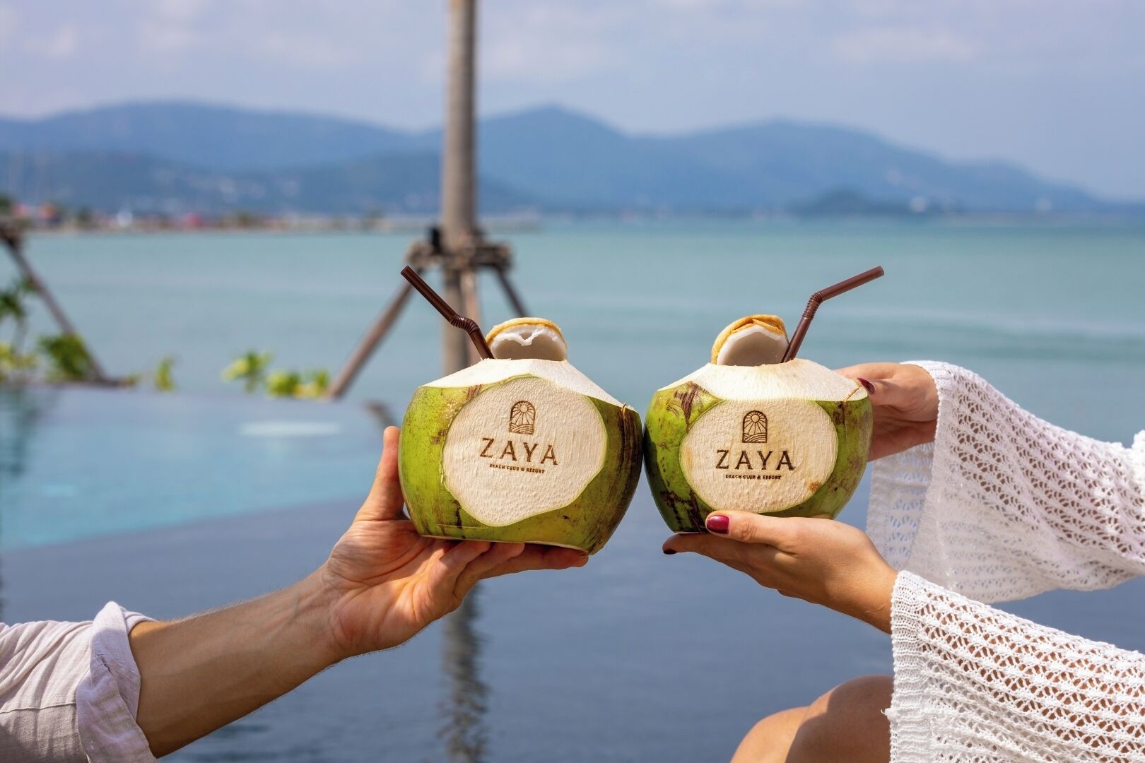 Two people holding a coconut drink with the brand name 'ZAYA' on it, with a body of water and mountains in the background.