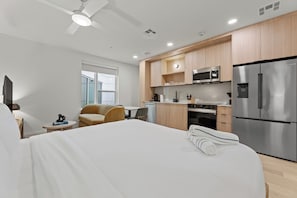A modern hotel room with a large bed, a small kitchenette, a ceiling fan, and a view of the city.