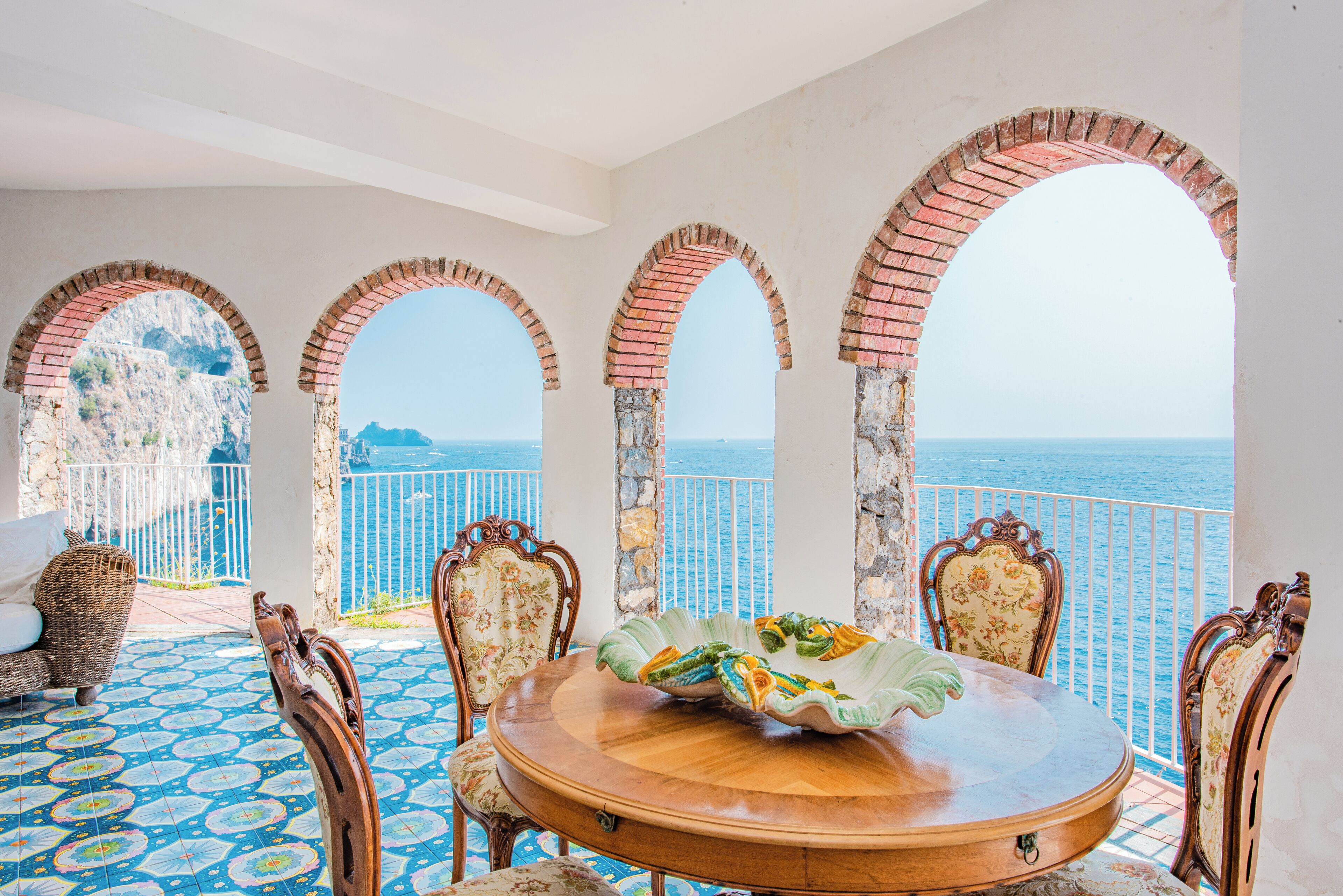 A dining area with a round table, chairs, and a view of the sea through arched windows.