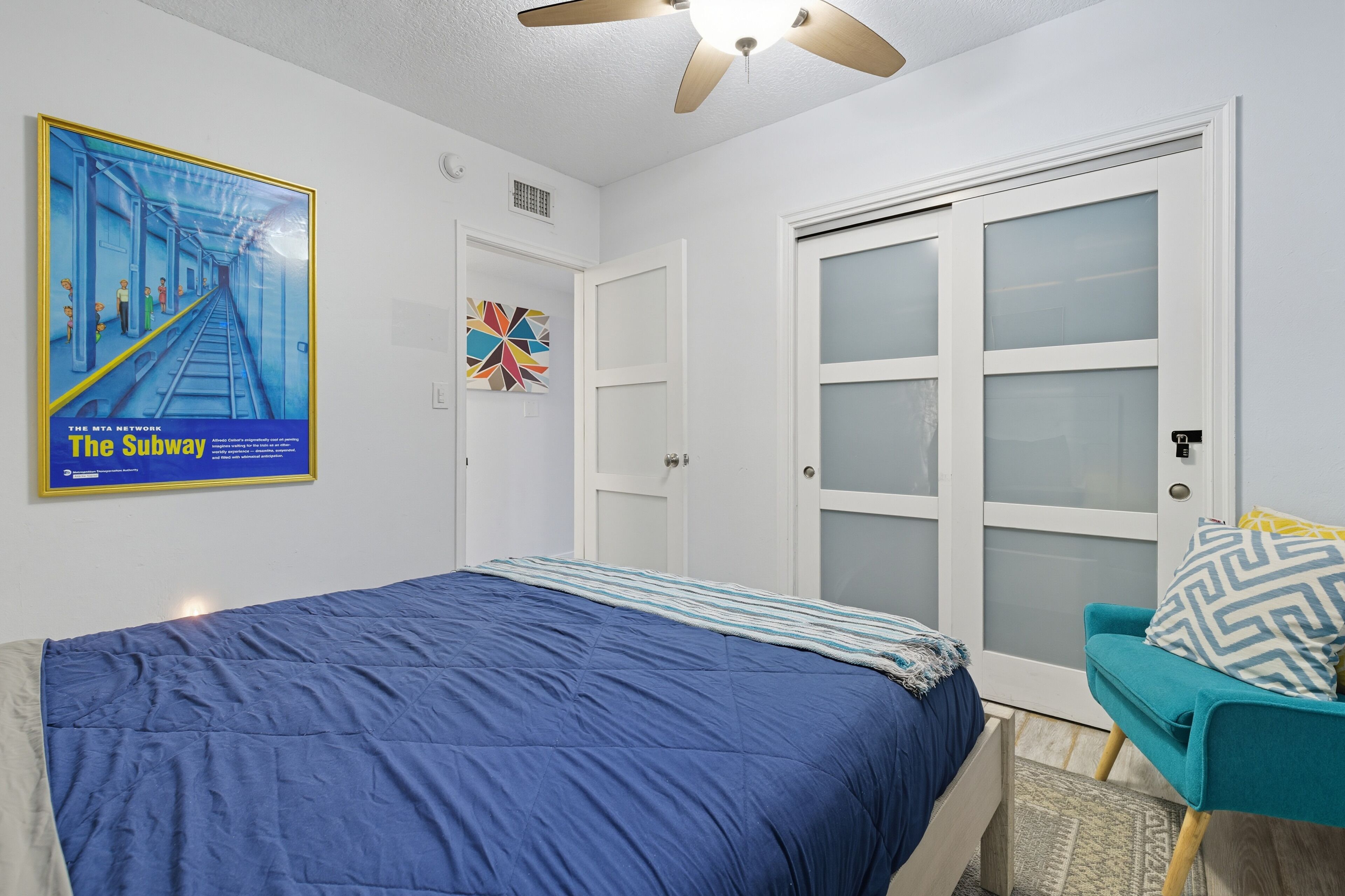 Una camera da letto con un letto, una sedia, una porta con pannelli trasparenti satinati, una ventola a soffitto e un poster appeso al muro.