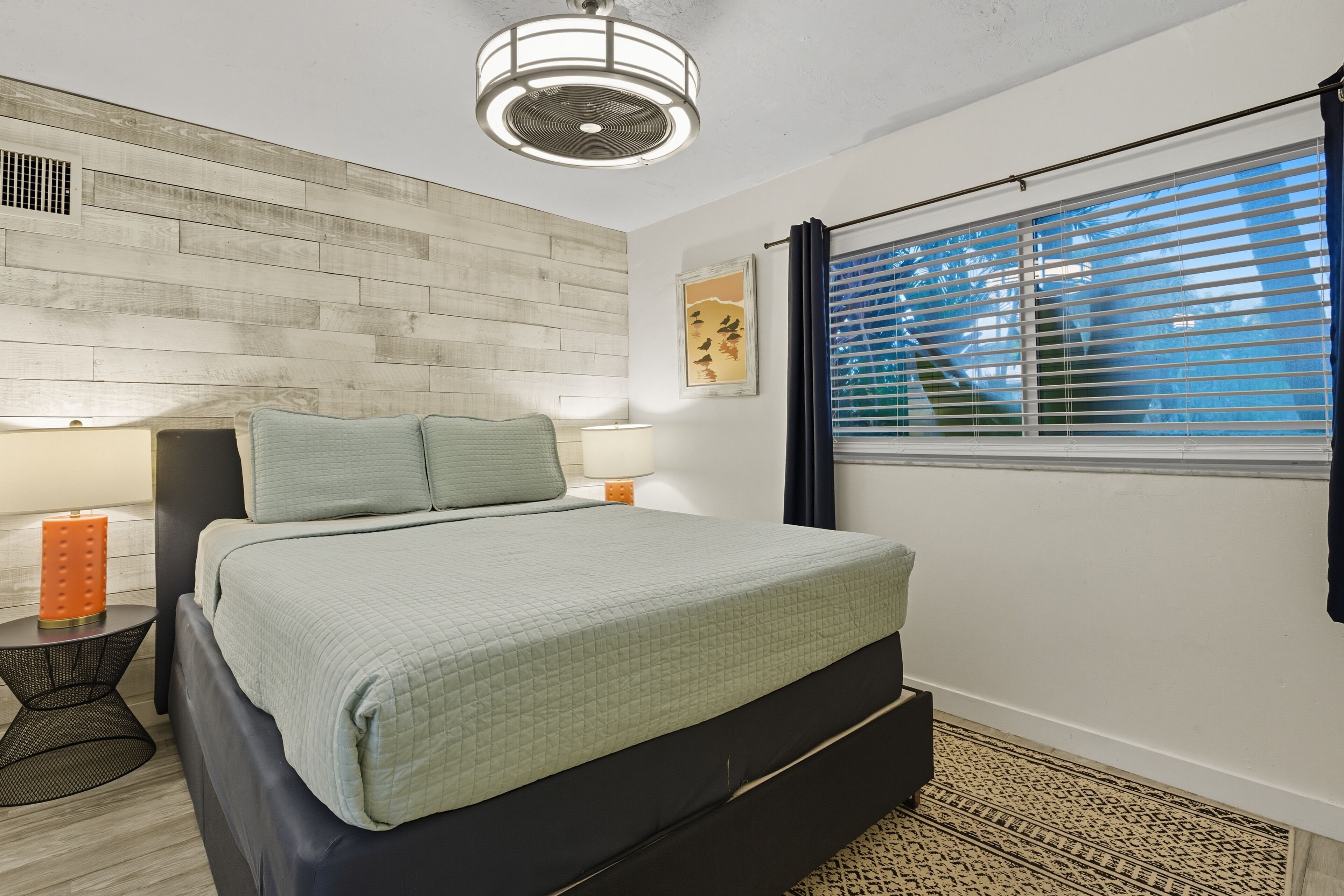 A modern bedroom with a bed, bedside lamps, a wall-mounted fan, and a window with blinds.