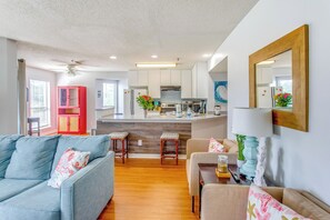 Ein modernes Wohnzimmer mit einem grauen Sofa, einem beigen Sessel und einem Holzessttisch mit zwei Stühlen. Im Hintergrund ist eine Küche mit weißen Schränken und einem roten Schrank erkennbar.