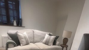 A living room with a beige sofa, patterned ottoman, and a small side table with a lamp. - Cosy cottage in the heart of Stow-on-the-Wold, pet-friendly (Stow-on-the-Wold)