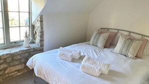 A neatly made bed with white linens and rolled towels, a stone wall, and a large window with natural light. - Cosy cottage in the heart of Stow-on-the-Wold, pet-friendly (Stow-on-the-Wold)
