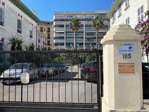 En hegnet parkeringsplads med flere biler, et skilt der angiver permanent adgangstilladelse for politi og nationalgarde, samt en bygning med altaner og palmer.