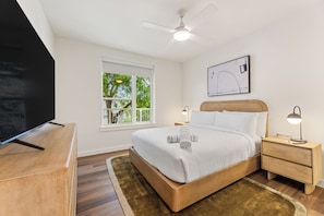 Une chambre à coucher comprenant un lit, des tables de chevet, un téléviseur, un ventilateur de plafond, une fenêtre et un tableau encadré au mur.