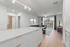 A modern kitchen with white cabinetry and a central island, a living area with a sofa and a coffee table, and a view through a glass door to an outdoor balcony.