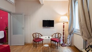 Une pièce avec un plancher en bois, une porte blanche, un téléviseur fixé au mur, une petite table ronde avec deux chaises et un lampadaire.
