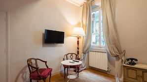 Une pièce avec un plancher en bois, un téléviseur fixé au mur, une table avec des chaises, une lampe, un radiateur et un téléphone posé sur une commode.