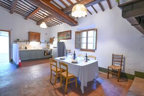 A dining area with a table set for four, wooden chairs, a kitchen in the background, and a ceiling with exposed beams.