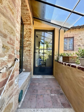 A narrow outdoor corridor with a glass door leading to a covered area, brick walls, and potted plants. - Apartment 'Tiziana' with Shared Garden and Wi-Fi (Vico dʼArbia)