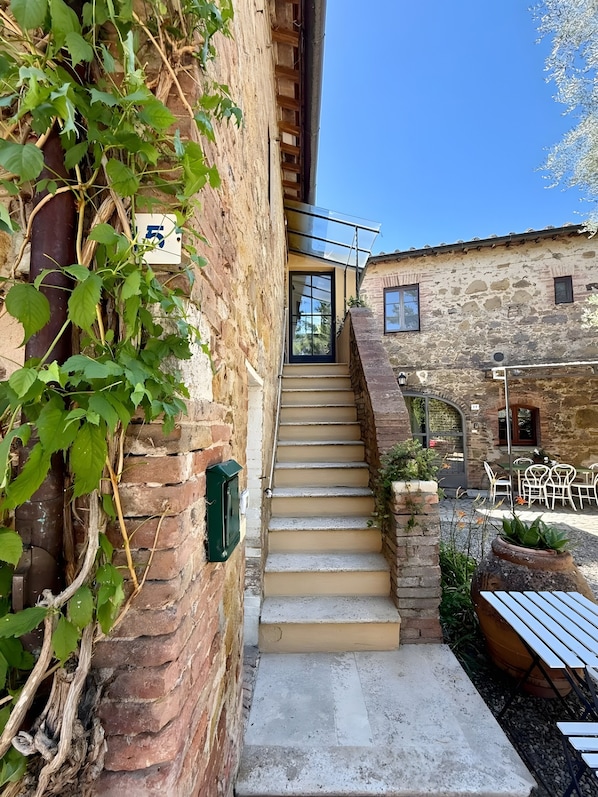 Un bâtiment en pierre avec un escalier menant à une porte, des sièges à l’extérieur et de la verdure.