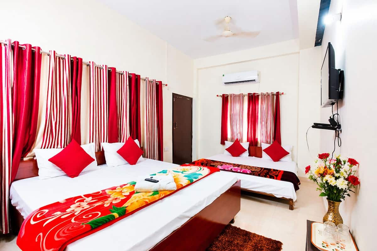 A hotel room with two beds, red and white curtains, a ceiling fan, an air conditioning unit, a TV, and a vase with flowers.