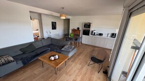 Een moderne woonkamer met een grijze hoekbank, een houten salontafel en een zwarte draadstoel. Achtergrond toont een keuken met witte kasten en apparatuur.