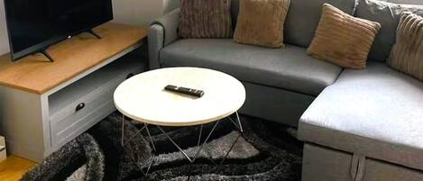 A living room with a grey sofa, a white coffee table, a TV on a wooden stand, and a black and grey rug.