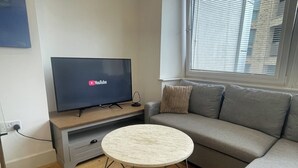 Una sala de estar moderna con un televisor de pantalla plana mostrando YouTube, un sofá gris, una mesa redonda de centro y una ventana con persianas.
