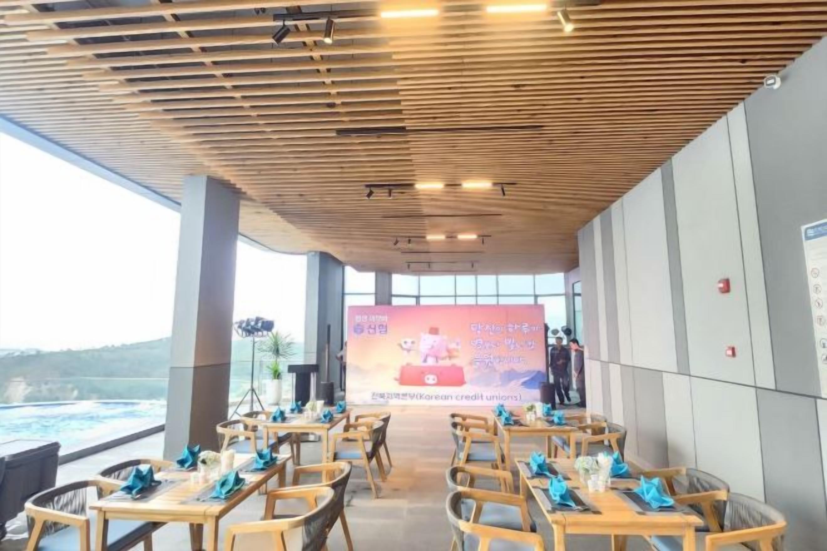 An indoor dining area with wooden tables and chairs, a large screen displaying an event, and a view of the outdoors.