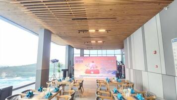 An indoor dining area with wooden tables and chairs, a large screen displaying an event, and a view of the outdoors.